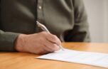 Senior man signing Last Will and Testament at wooden table indoors, closeup