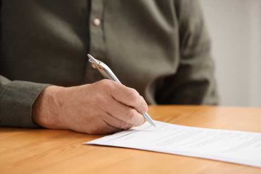 Senior man signing Last Will and Testament at wooden table indoors, closeup